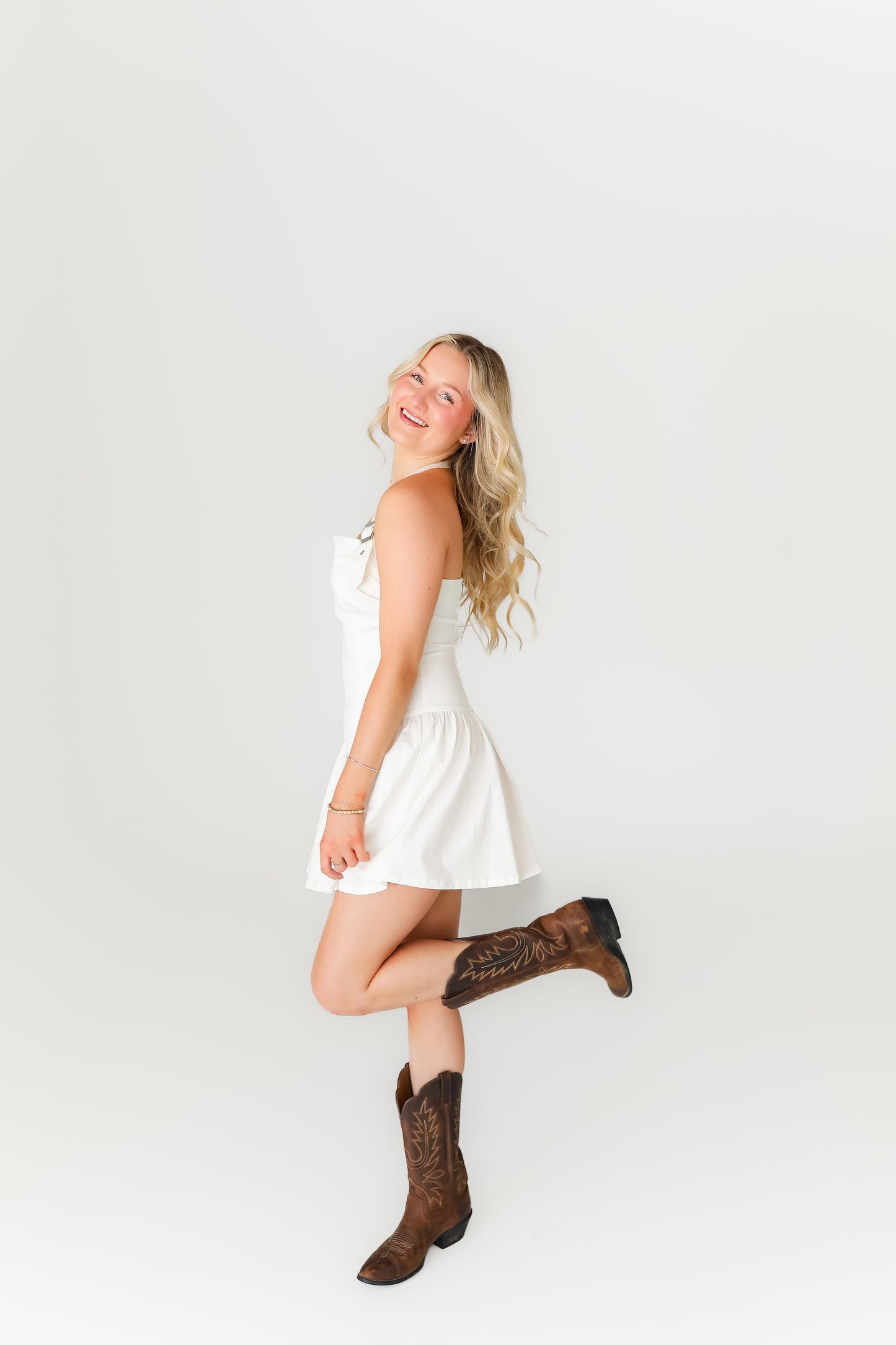 Woman in a white dress and brown boots posing against a plain background