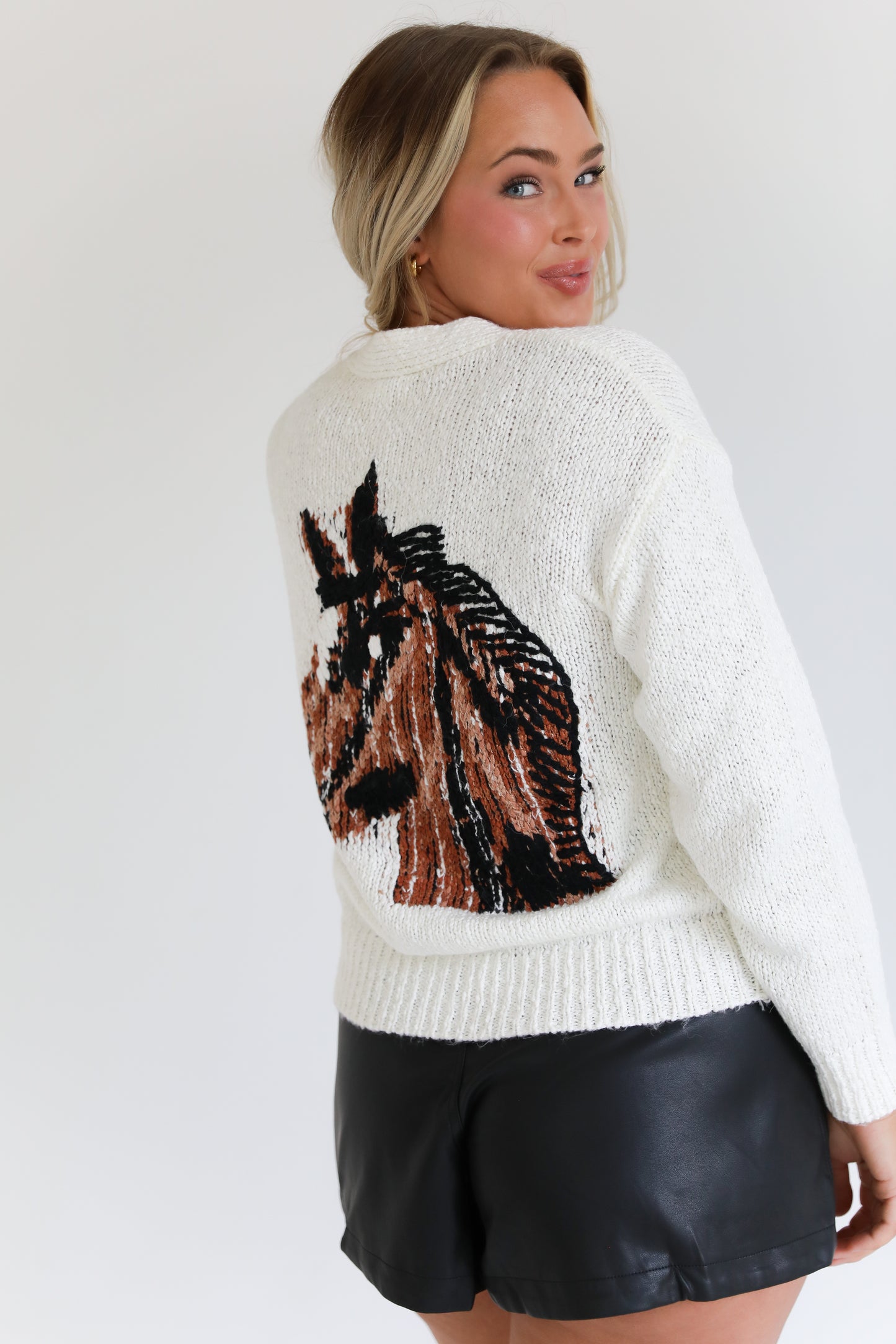 Woman wearing a white knit sweater with a horse design on the back & embroidered horseshoe on the front